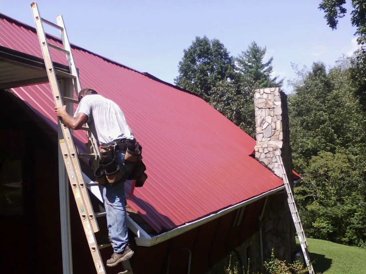 Roofer on a ladder beside a red metal roof and stone chimney — Soffit & Fascia Repair by Roofing Brothers in Allred